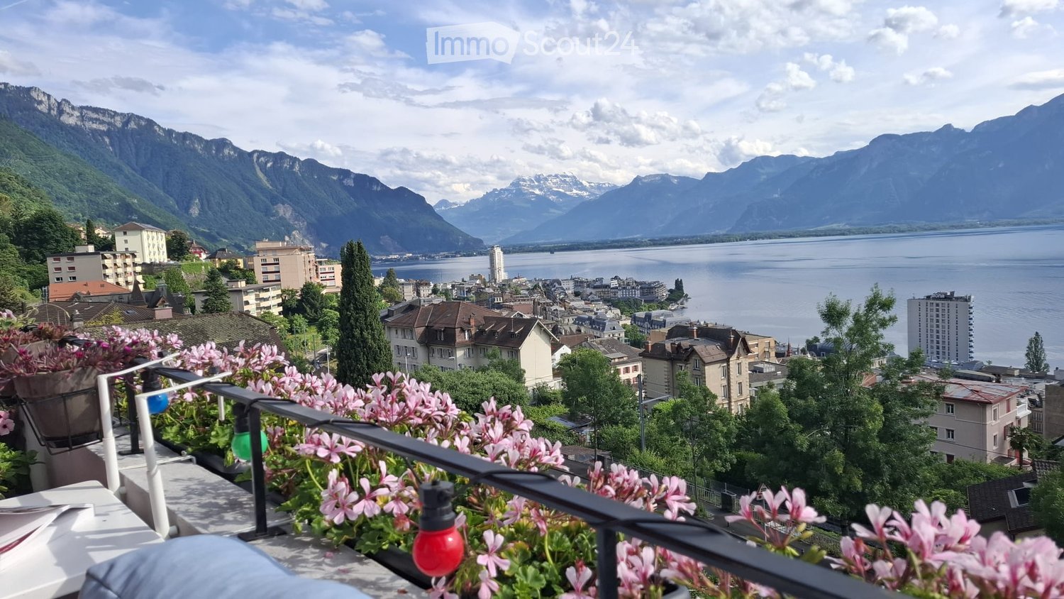 Balcony with pink flowers, overlooking a cityscape with mountains and a lake in the background.