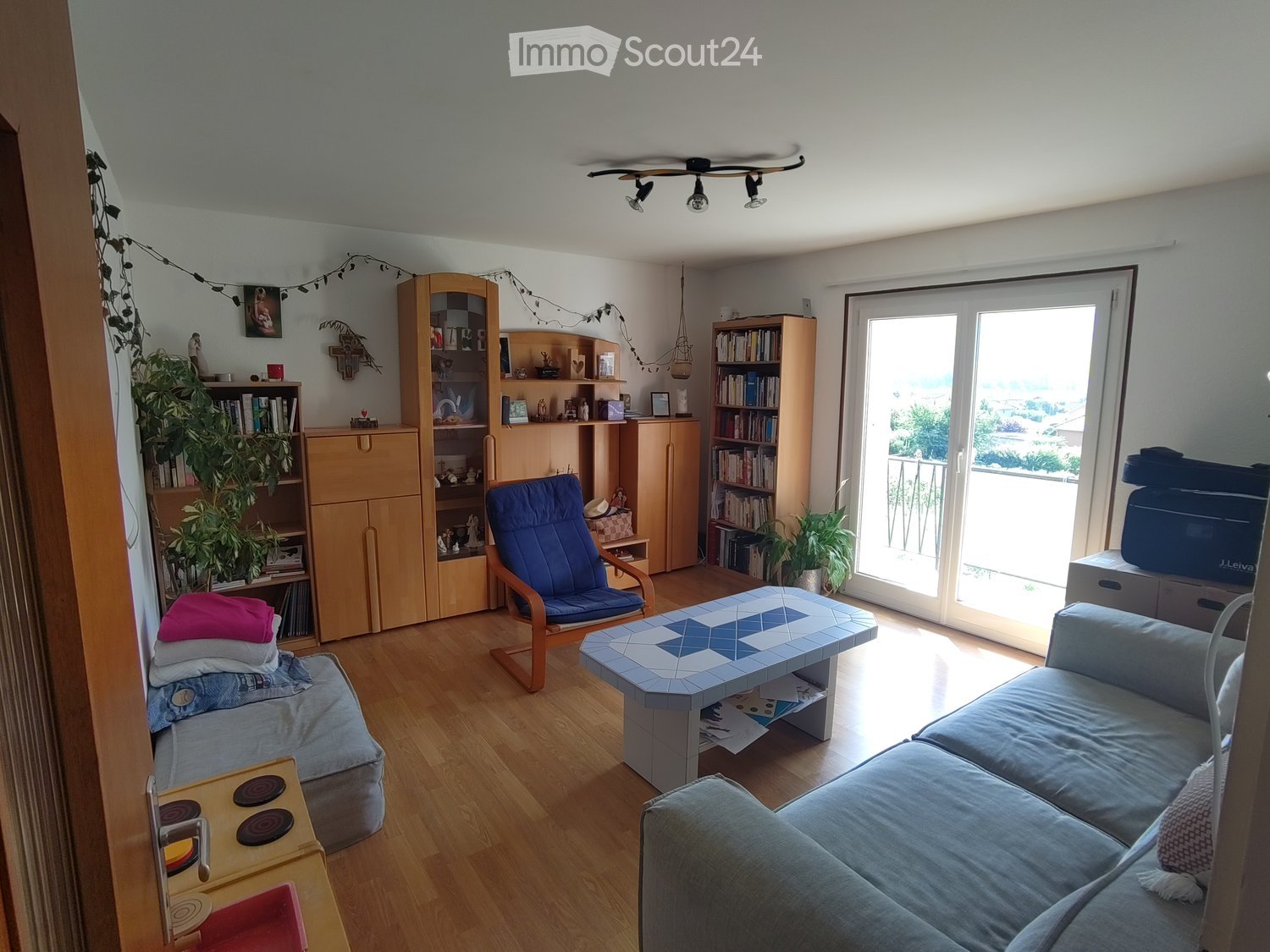 Living room with wooden flooring, a grey sofa, a blue armchair, a small table with a blue and white patterned top, bookshelves, and a balcony door leading outside.