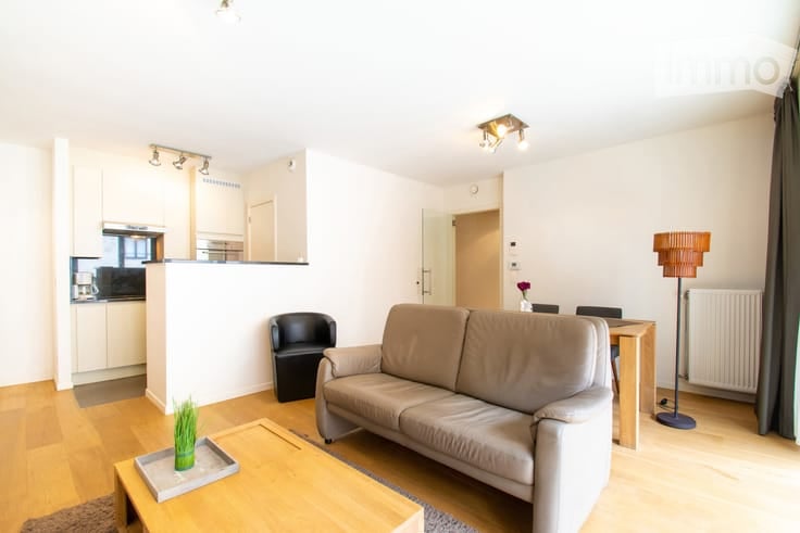 Modern, open-plan living and dining area with light wood flooring, white walls, and contemporary furniture including a sofa, coffee table, dining table, and chairs. A small kitchen area is visible in the background with cabinets and a microwave.