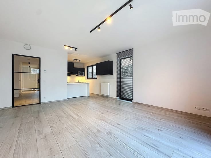Open-plan unfurnished room with light wooden flooring, white walls, and a modern kitchen area in the background. The kitchen features black cabinets, a white countertop, and built-in appliances.