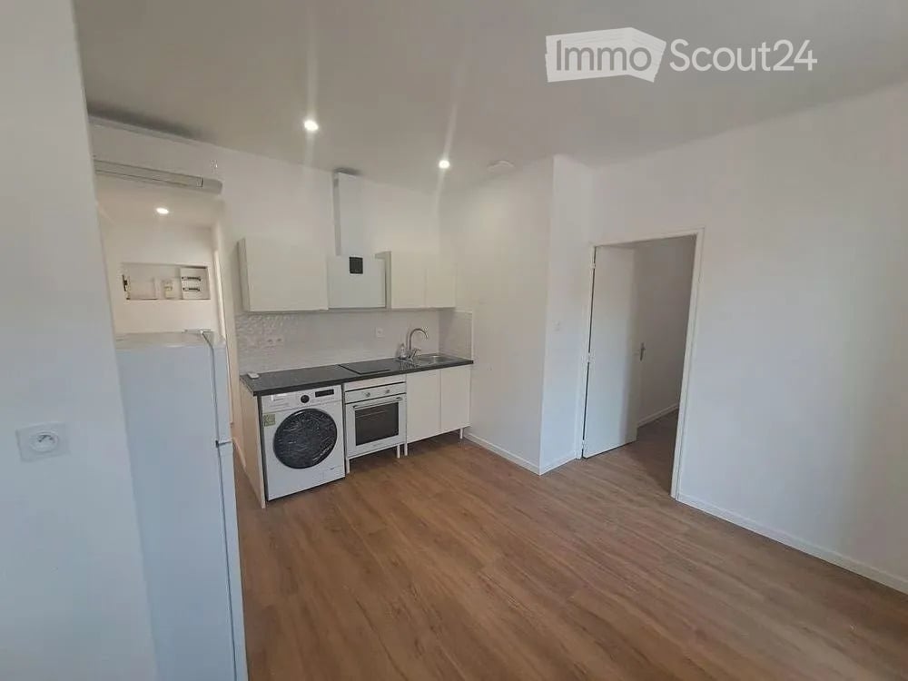 A modern, compact kitchen with white cabinets, a black countertop, and integrated appliances including a refrigerator, washing machine, and oven. The space features wooden flooring and a small sink area with a faucet.
