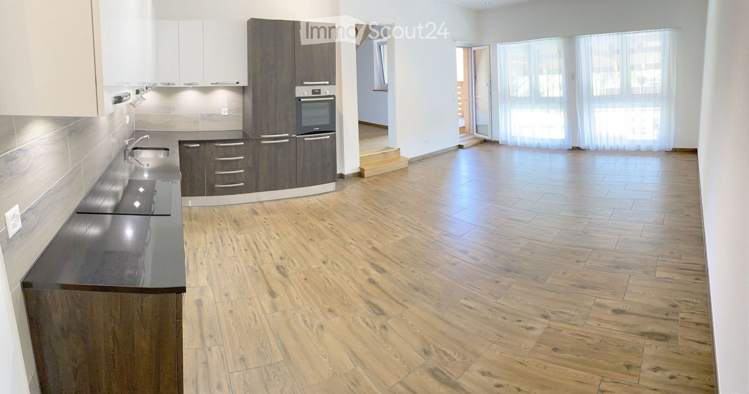 Modern kitchen with wooden flooring, dark grey cabinets, stainless steel sink, and built-in oven. Open space with large windows and a view to another room.