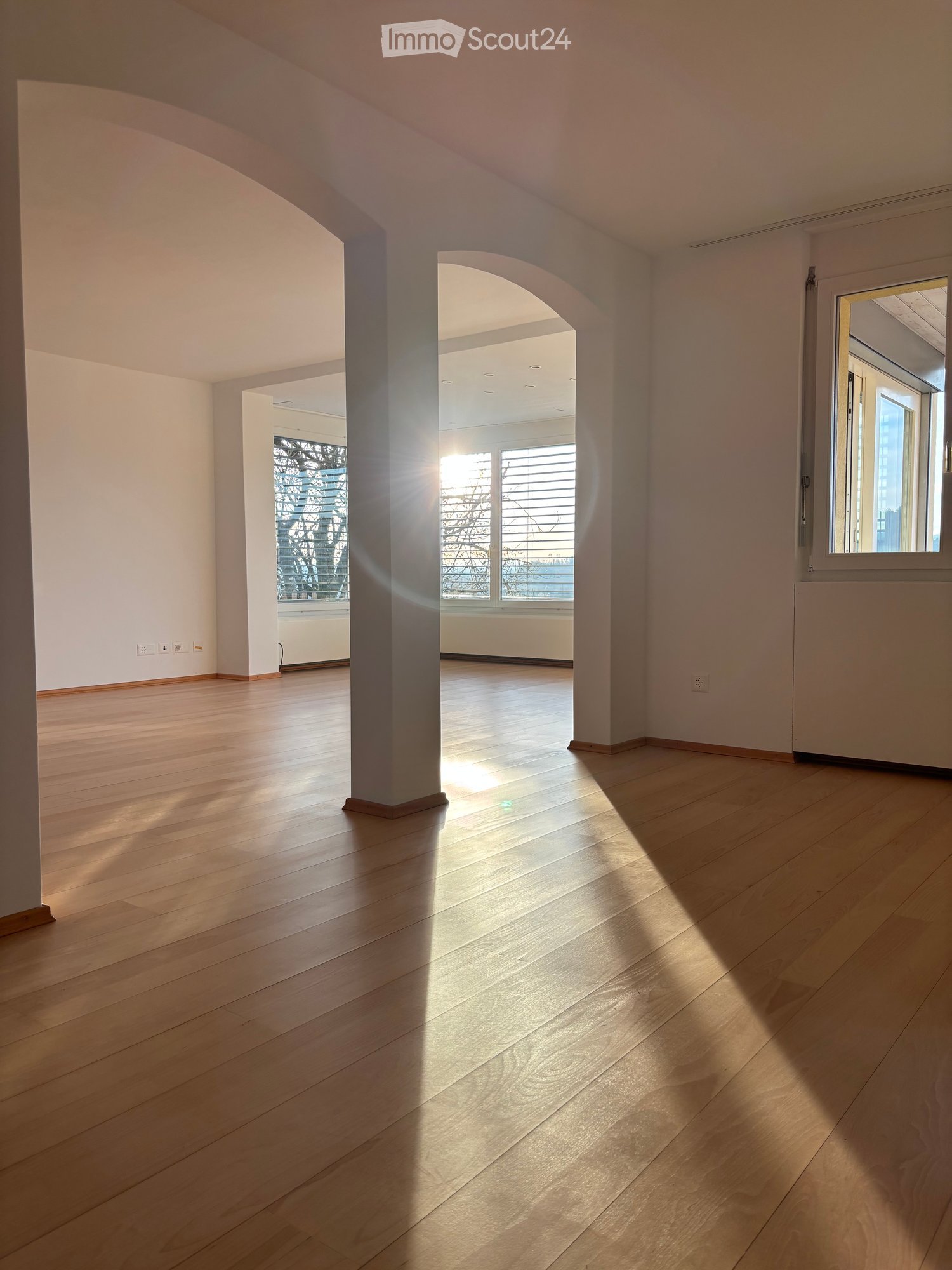 Empty room with wooden flooring, white walls, and large windows allowing natural light to enter. The space is open and bright with visible architectural columns and electrical outlets.
