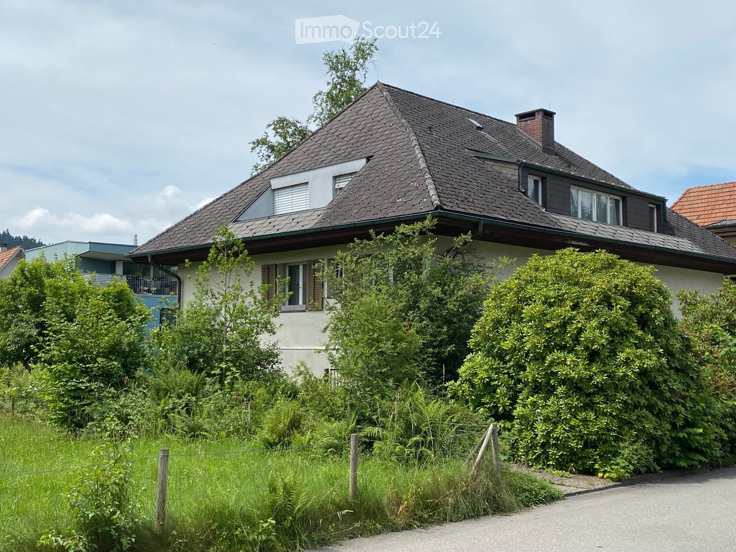 Two-story house with a dark tiled roof, multiple windows, and a chimney. The house is surrounded by greenery and has a fenced front yard.
