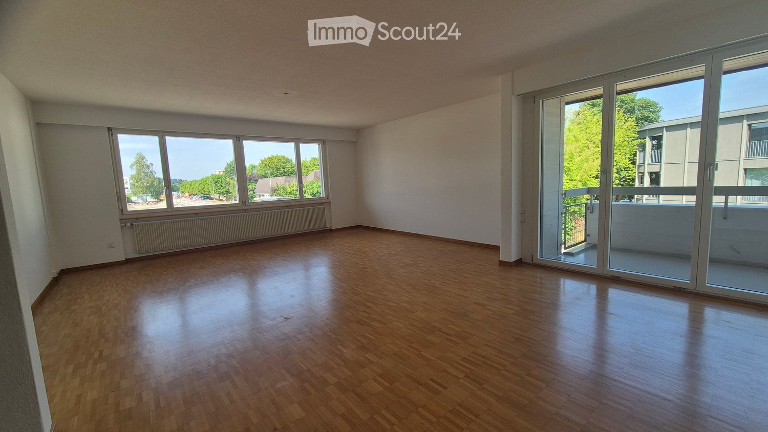 Empty room with wooden flooring, large windows, and sliding glass doors leading to a balcony.