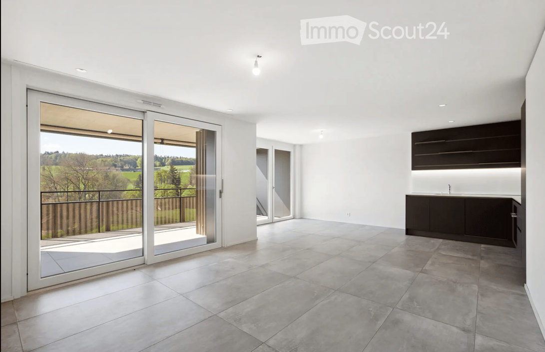 Bright, open-plan space with large sliding glass doors leading to a balcony, modern kitchen area with dark cabinets, and a view of a green landscape.