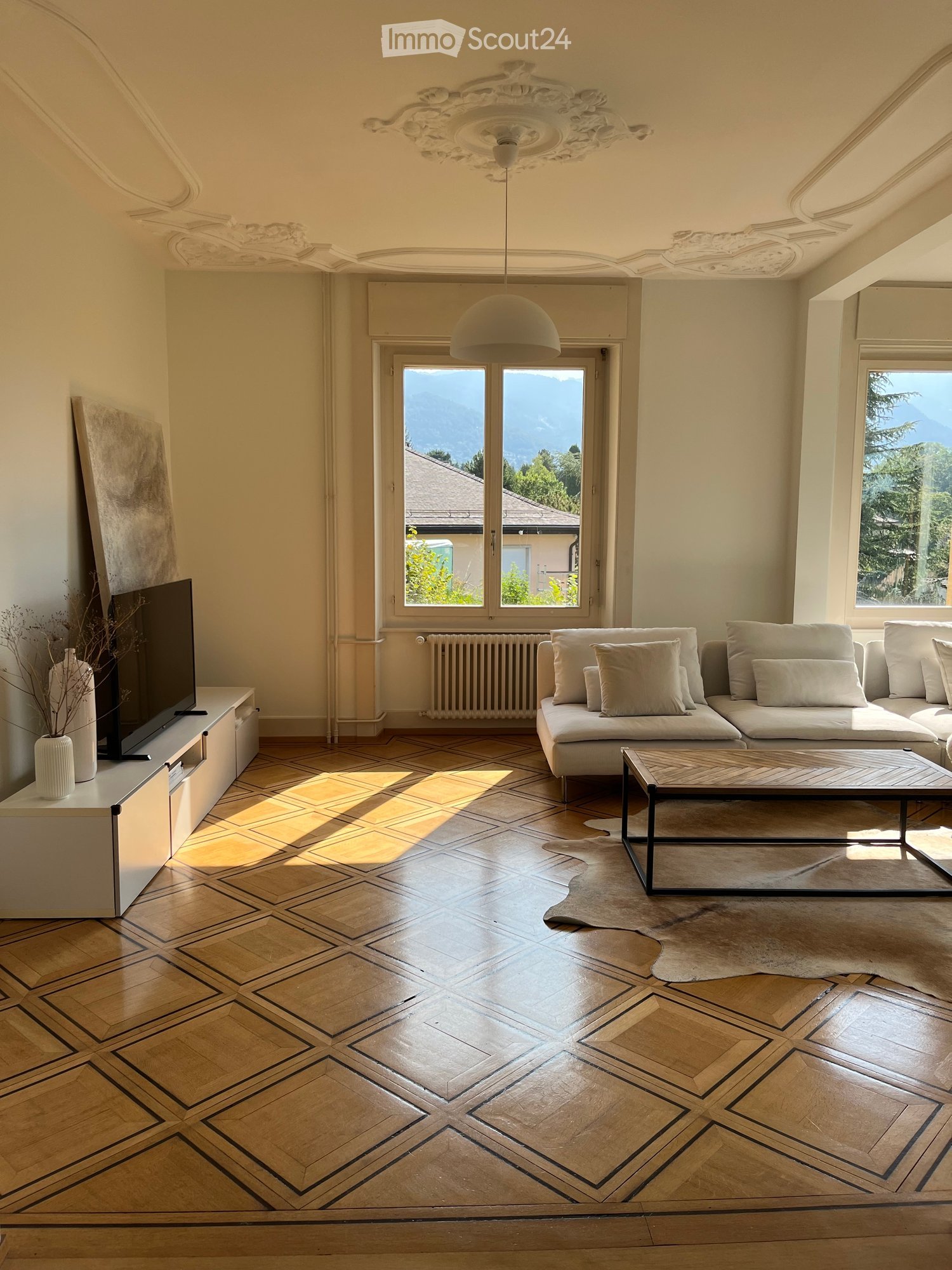 Elegant living room with a geometric parquet floor, large windows offering a scenic mountain view, a modern sofa set, a sleek coffee table, and a TV stand with a screen.