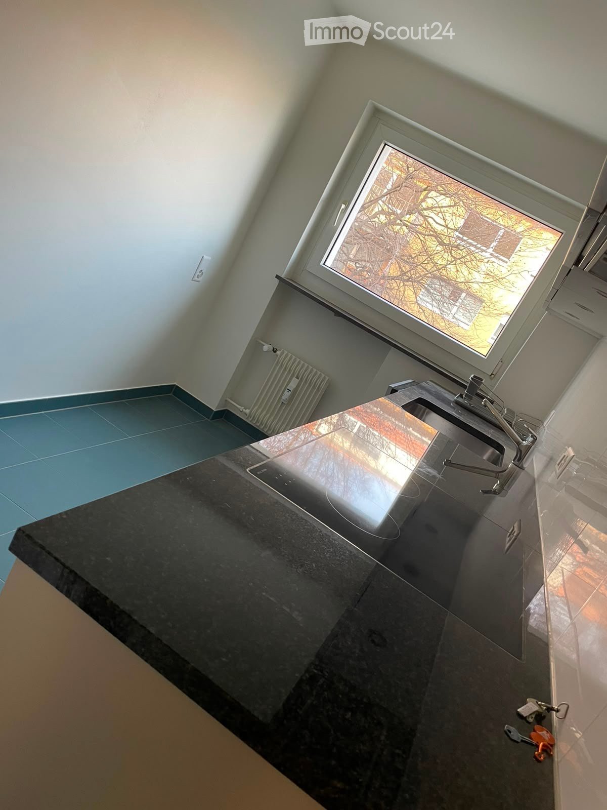 Modern kitchen with black granite countertop, stainless steel sink, and a window above the sink. The floor is tiled in blue and black, and the walls are white with a radiator visible.