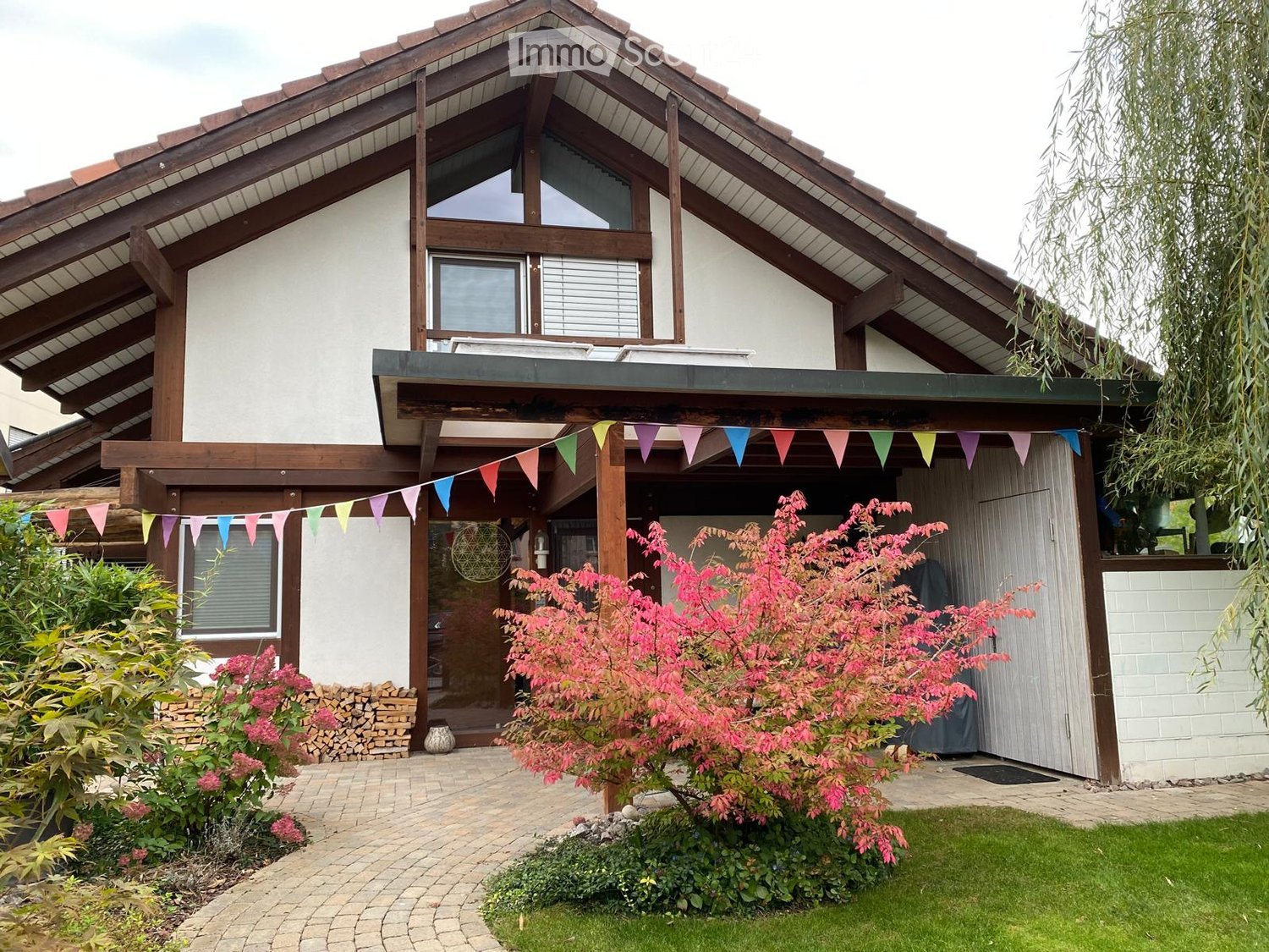 Two-story house with a wooden roof and white walls, featuring a covered entrance with colorful bunting, a well-maintained garden with pink flowering shrubs, and a neatly paved pathway.
