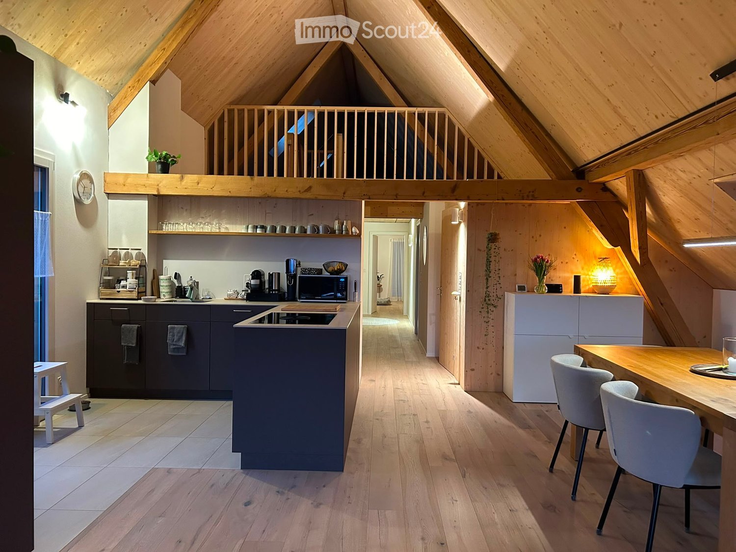 Modern kitchen with dark grey cabinets, wooden countertops, and a dining area with white chairs and a wooden table. The space features exposed wooden beams and a lofted sleeping area above.