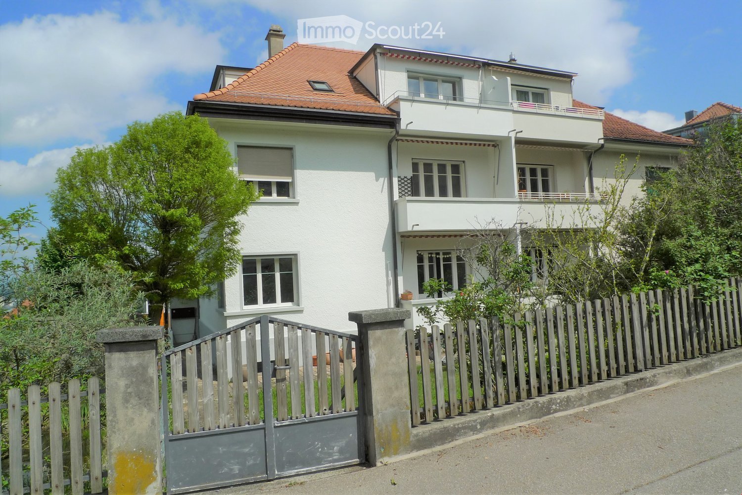 Two-story white house with red-tiled roof, multiple windows, and a balcony on the upper floor. The house is surrounded by a wooden fence and greenery.