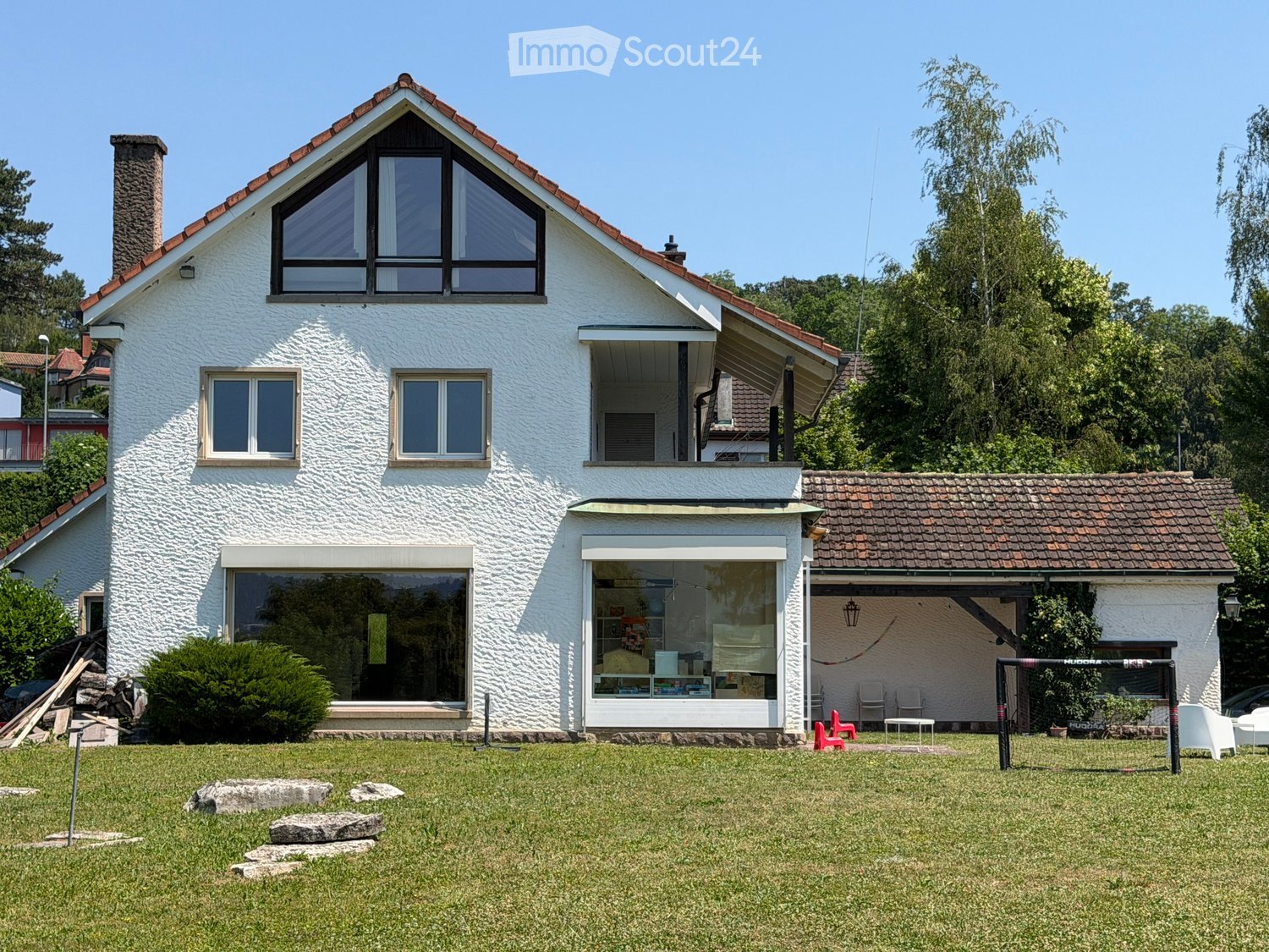 Two-story white house with red-tiled roof, large windows, and a balcony. The house is surrounded by a well-maintained lawn with scattered rocks and a small outdoor seating area with chairs and a table.