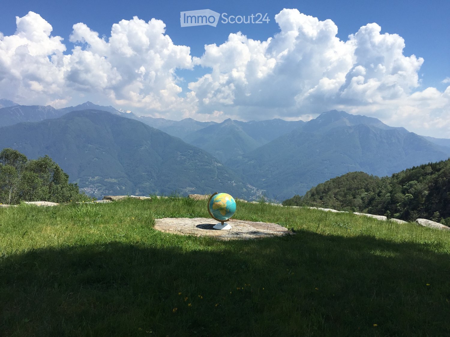 A grassy hilltop with a globe on a stone platform, overlooking a mountain range under a partly cloudy sky.