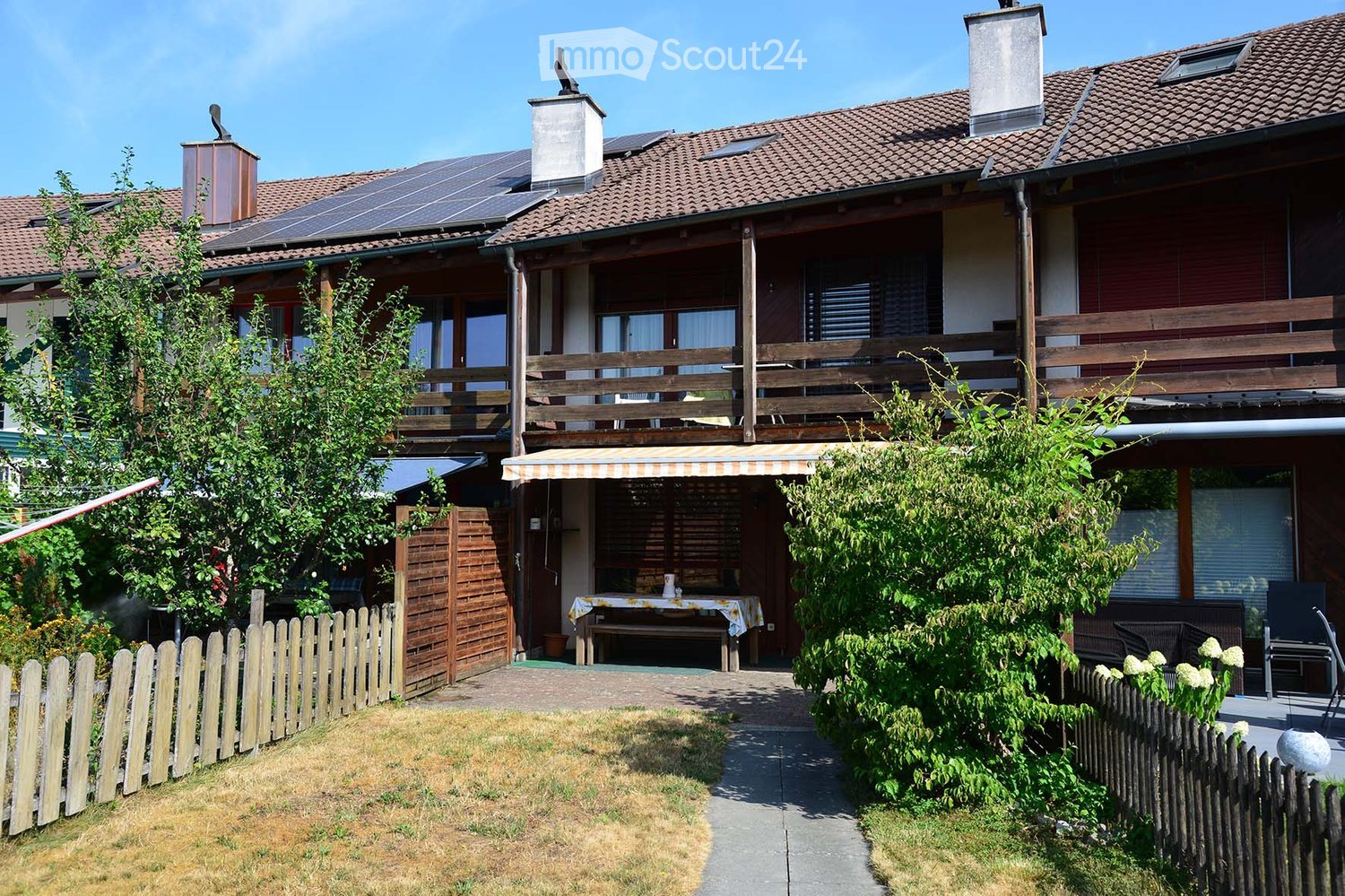 Two-story house with a tiled roof, solar panels, and a wooden balcony. The front yard includes a wooden fence, a small patio with a table and chairs, and lush greenery.