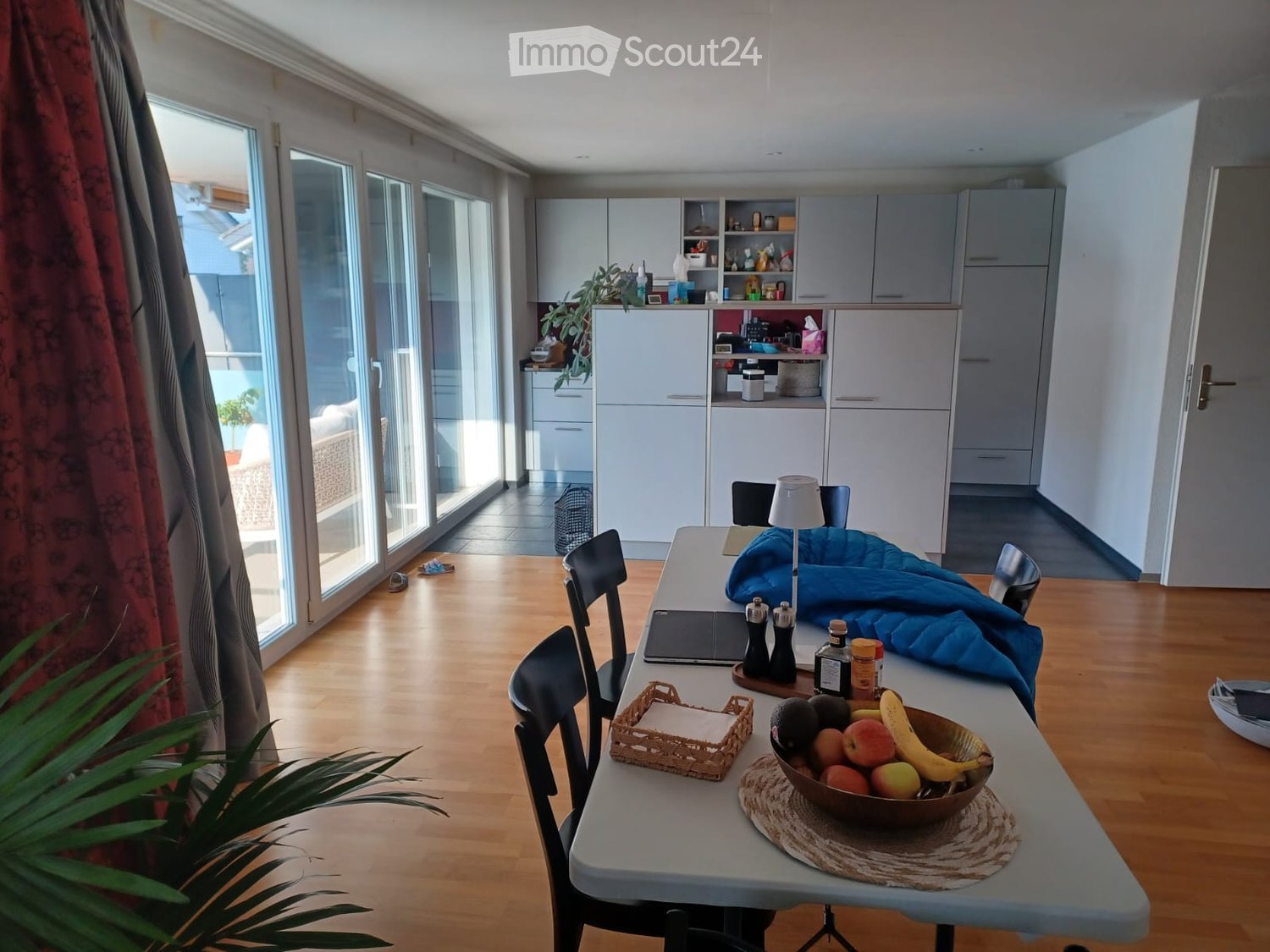Bright, modern kitchen and dining area with white cabinets, wooden flooring, and large windows. A dining table with chairs is set with a bowl of fruit, a laptop, and a lamp. The kitchen features open shelving with various items and a small plant on the co