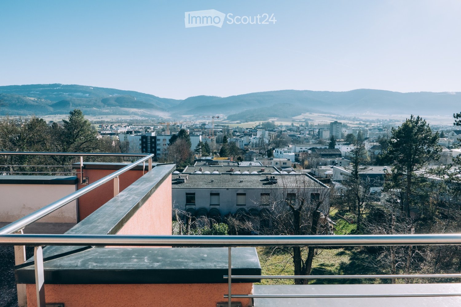 Balcony with metal railing overlooking a cityscape, with buildings, trees, and distant hills visible.