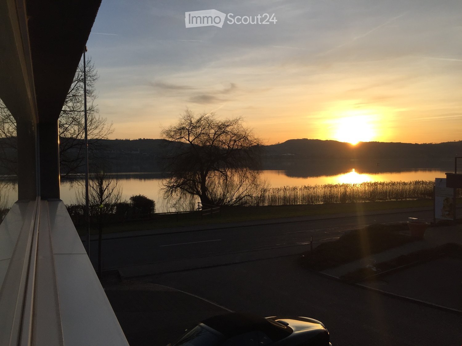 Balcony overlooking a serene lake at sunset, with trees and distant hills visible.