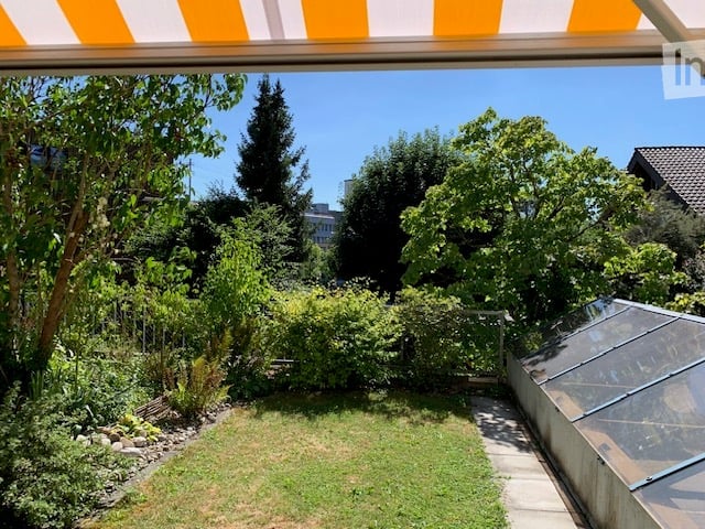 Well-maintained garden with a lawn, various trees, shrubs, and a small greenhouse. The area is enclosed and features a clear, sunny sky.