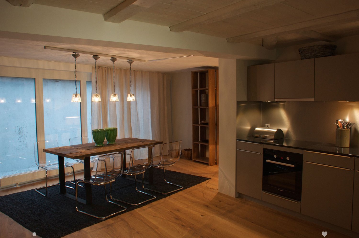Modern open-plan kitchen and dining area with wooden flooring, pendant lights, a wooden dining table with four transparent chairs, and a grey kitchen with built-in appliances.