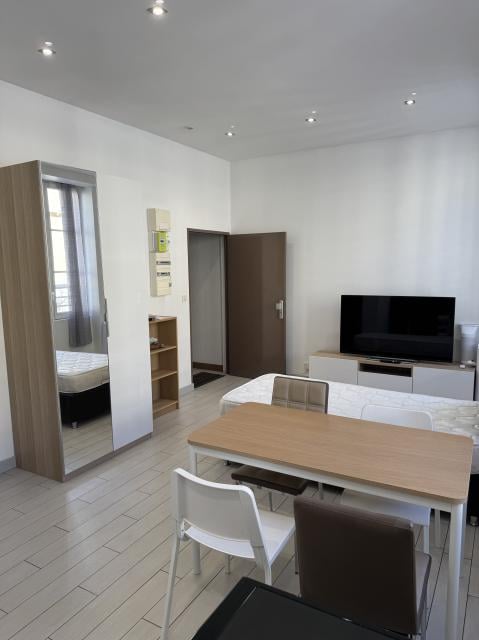 A modern, well-lit room with a large mirror, a wooden table with chairs, a TV on a stand, and a shelf with various items.
