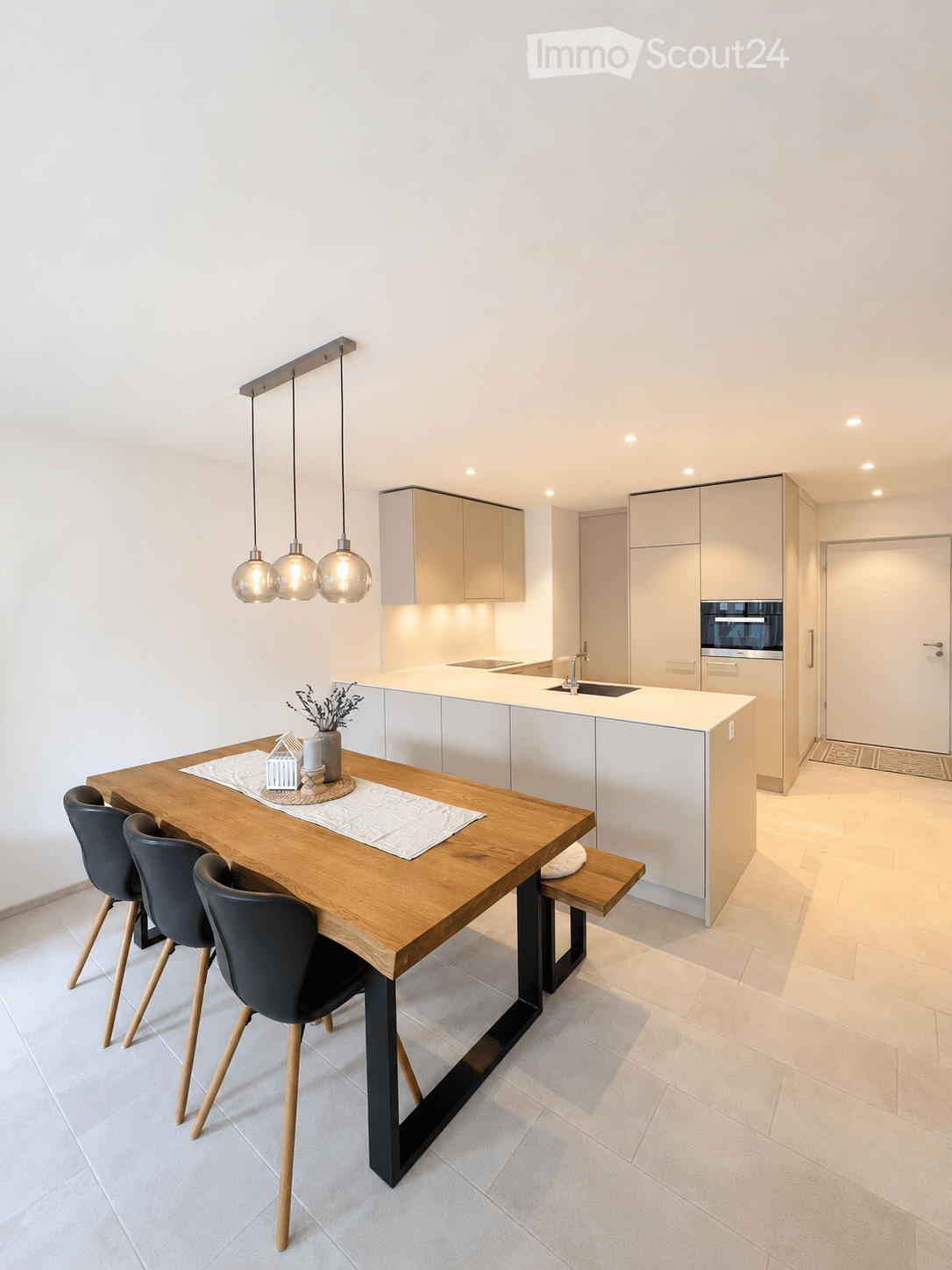 Modern kitchen with white cabinets, a wooden dining table, four black chairs, pendant lights, and a sink with a faucet. A vase with dried flowers and a candle are placed on the table.
