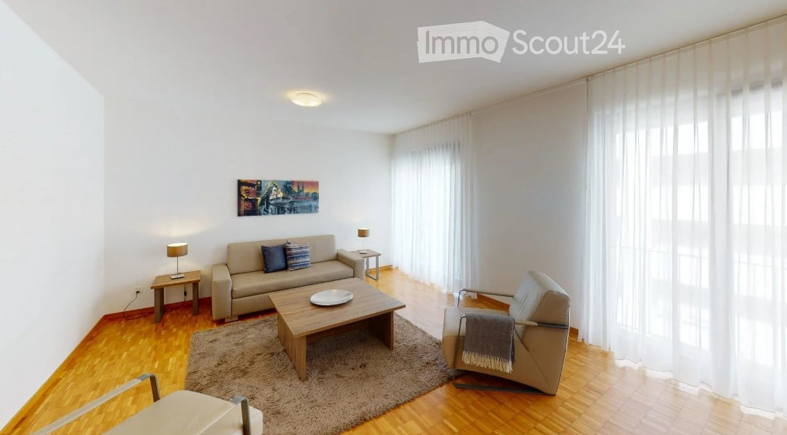 Bright living room with wooden flooring, large windows with white curtains, a beige sofa with two throw pillows, a wooden coffee table with a decorative plate, an armchair with a throw blanket, side tables with lamps, and a picture frame on the wall.