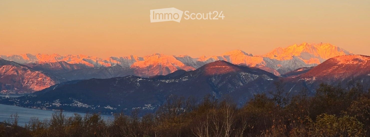 Panoramic view of snow-capped mountains at sunset, with a lake in the foreground and trees in the middle ground.
