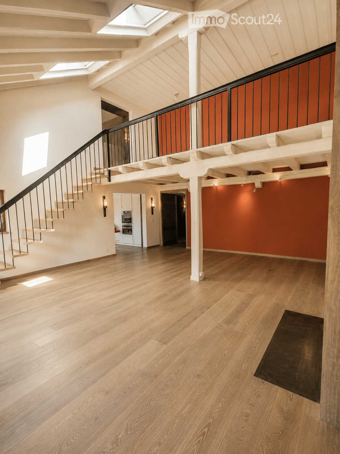 Spacious hallway with wooden flooring, white walls, and a staircase leading to an upper level. Features a black railing, warm lighting, and a glimpse of a kitchen area with white cabinets and stainless steel appliances.