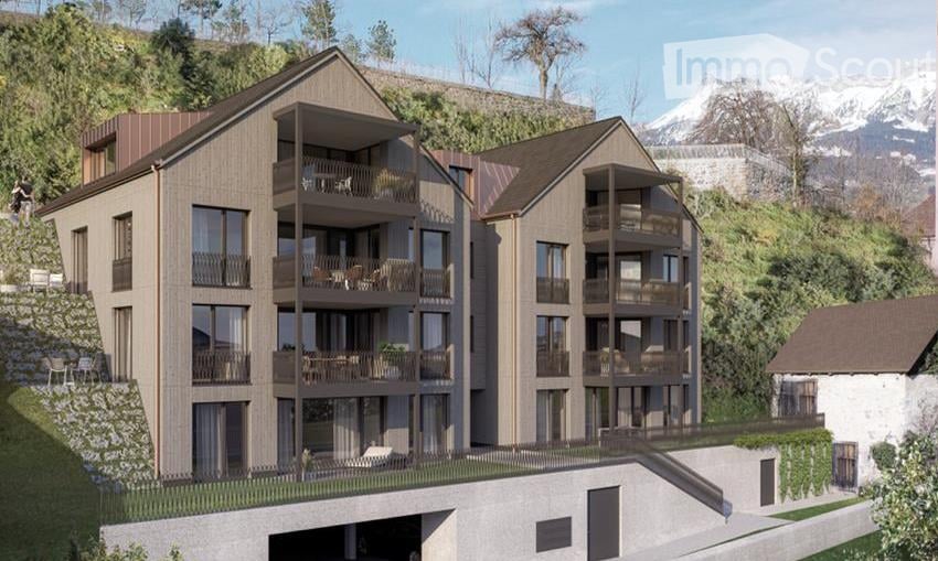 Modern multi-story residential building with balconies, large windows, and a raised ground floor. The building features a contemporary design with wooden and concrete elements, situated in a scenic, elevated location with a view of mountains in the backgr