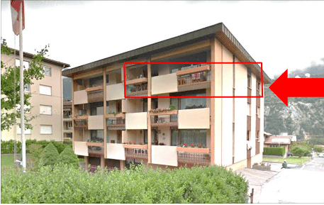 Multi-story apartment building with balconies and large windows, surrounded by greenery and mountains.