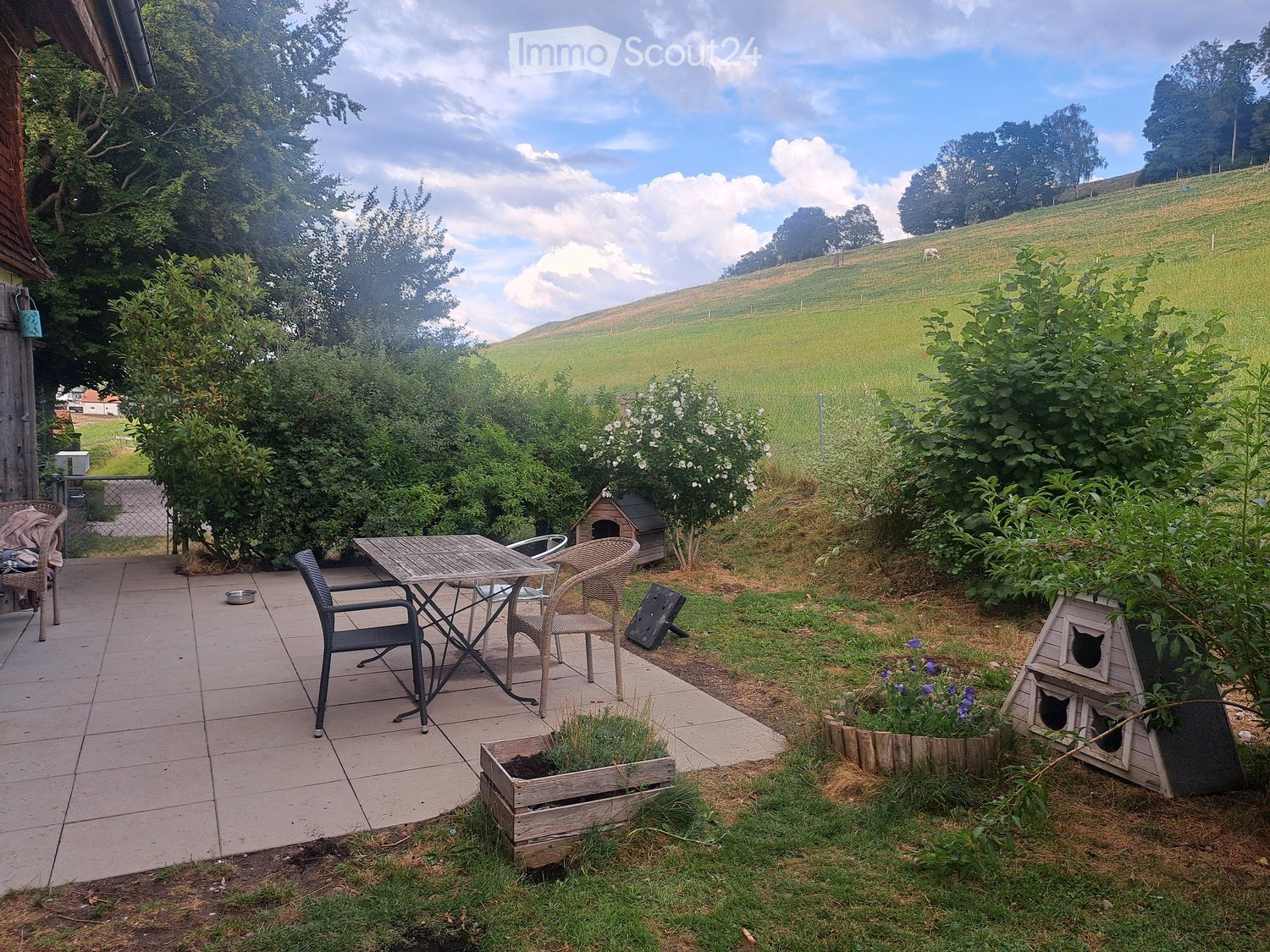 Garden area with tiled patio, outdoor table and chairs, cat house, and flower pots.