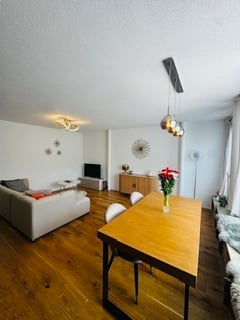 Bright, modern living and dining area with wooden flooring, white walls, and contemporary furniture including a sofa, coffee table, dining table, and chairs.