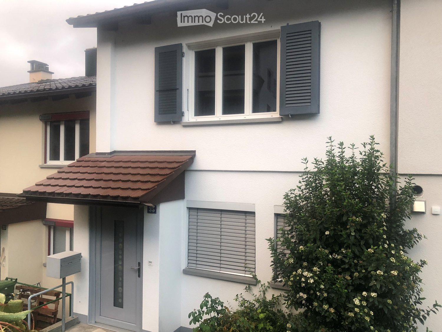 Two-story house with white walls, grey shutters, and a tiled roof. Features include a small balcony, a mailbox, and a small garden with shrubs and flowers.