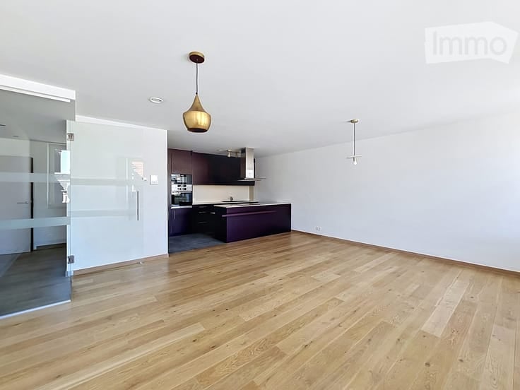 Empty room with wooden flooring, white walls, and a modern kitchen area visible in the background. No furniture or equipment present.