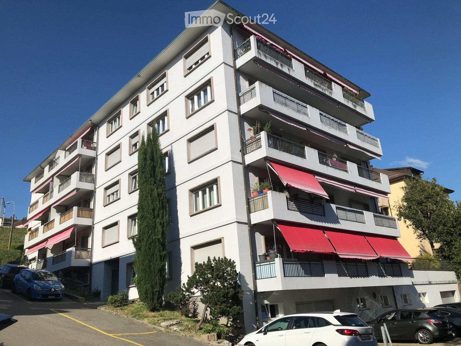 Multi-story residential building with balconies, red awnings, and surrounding greenery.