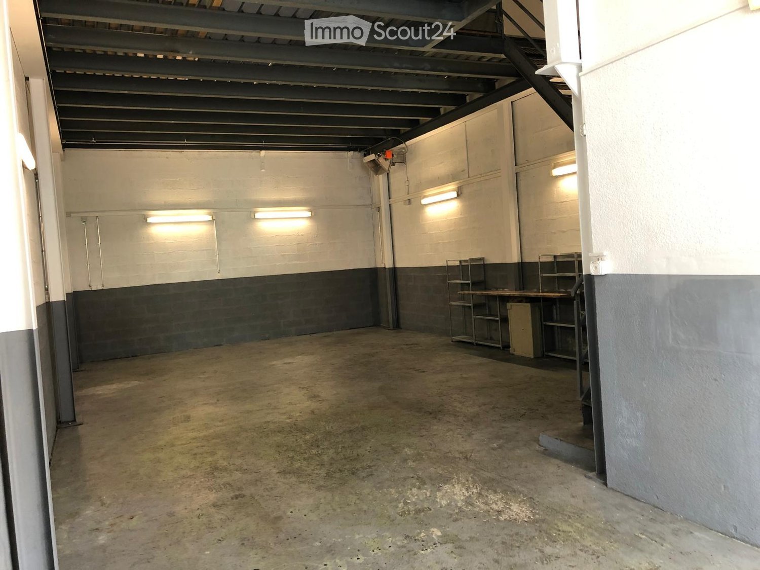 Empty room with concrete floor, white and gray walls, metal shelving units, and a staircase leading upwards.