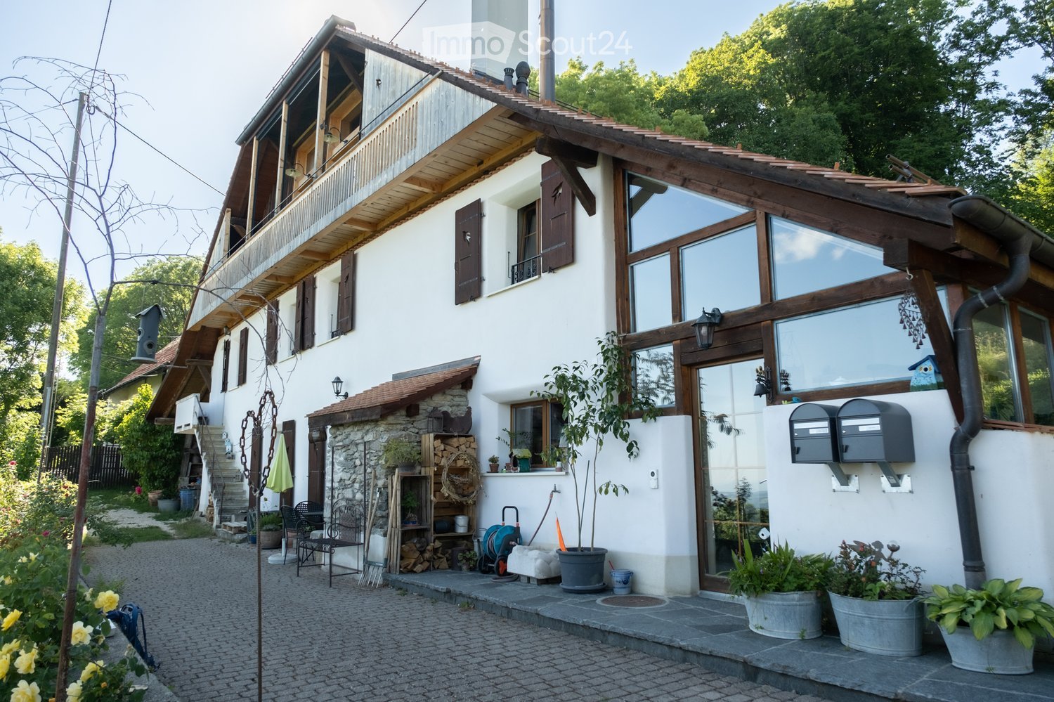 Two-story house with white walls, wooden accents, large windows, and a balcony on the upper level. The house features a stone chimney, a small garden area with potted plants, and a paved driveway.