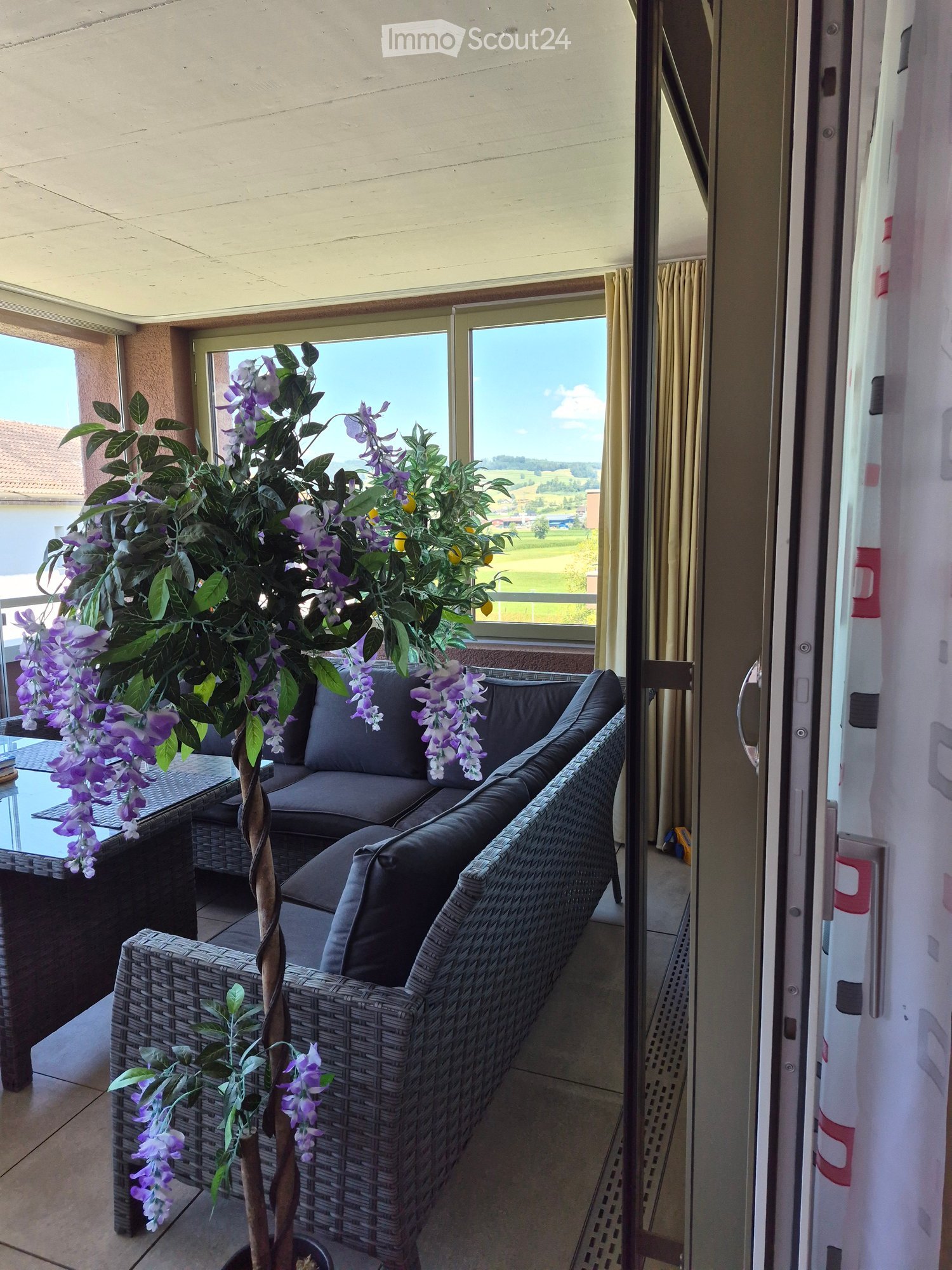 Balcony with a wicker sofa, glass table, and decorative plant with purple flowers, overlooking a scenic countryside view.