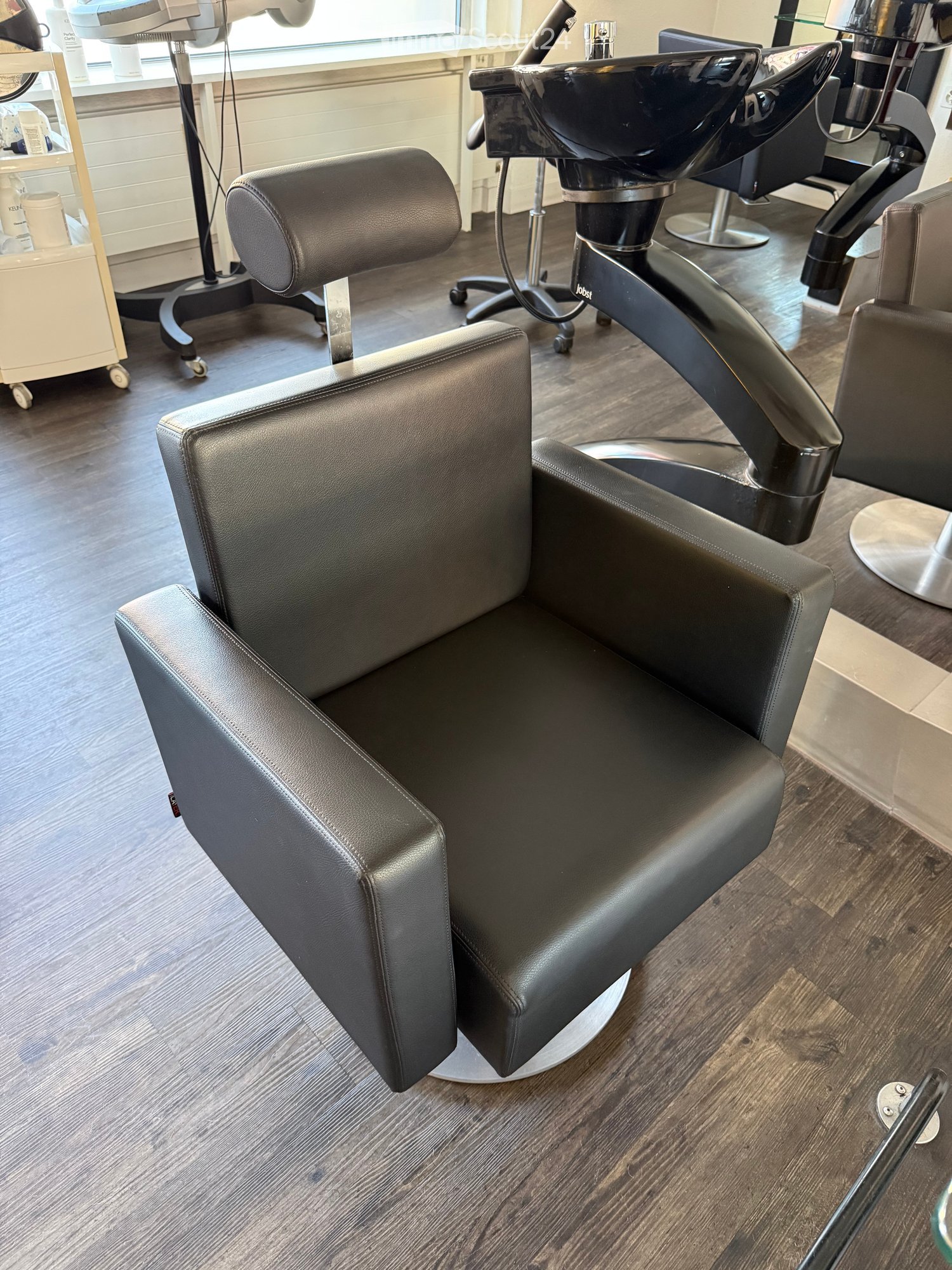 Modern salon with black leather chairs, a sink, and a stool, set on a wooden floor.