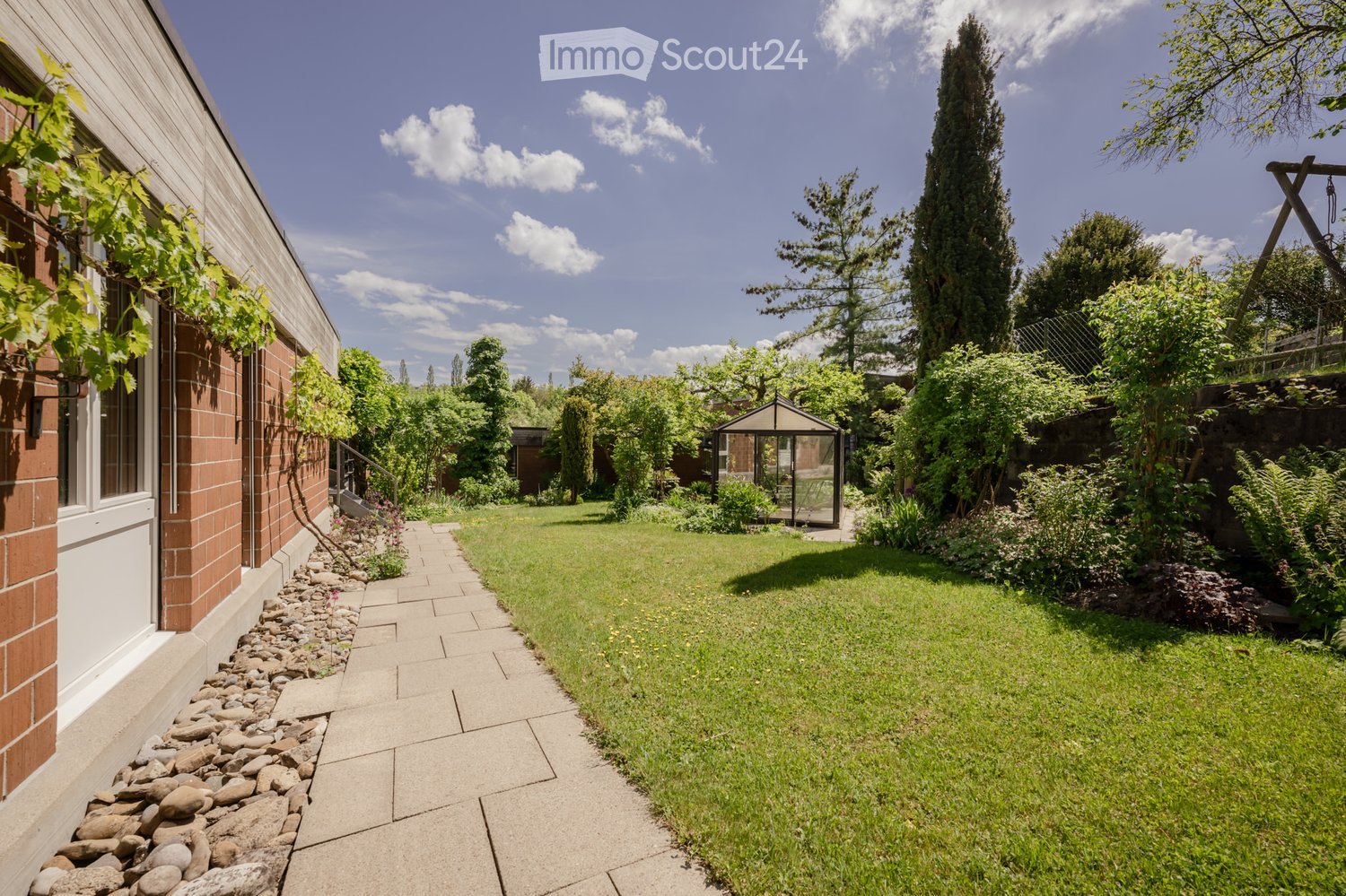 A well-maintained garden with a paved pathway, grassy lawn, and various trees and shrubs. A small gazebo is visible in the background, and the garden is bordered by a brick wall and a wooden fence.