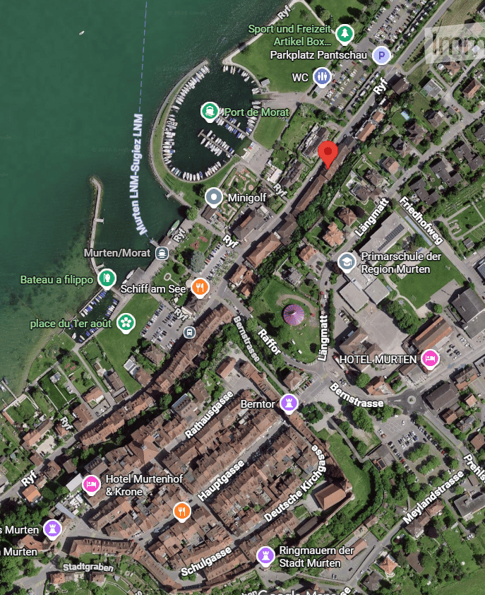 Aerial view of a town with buildings, streets, and a waterfront area with a marina. Visible landmarks include a hotel, school, and recreational areas.