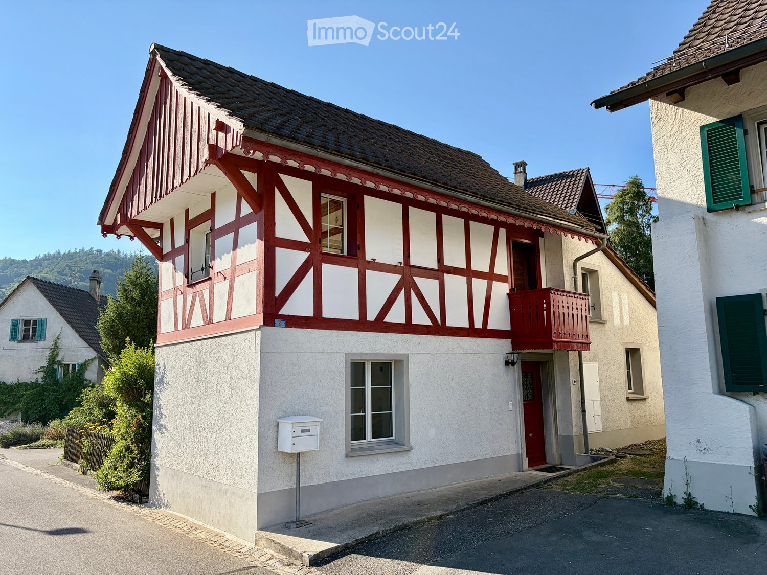 Two-story house with red and white timber framing, red shutters, and a small balcony. The house has a tiled roof and is surrounded by greenery and neighboring houses.