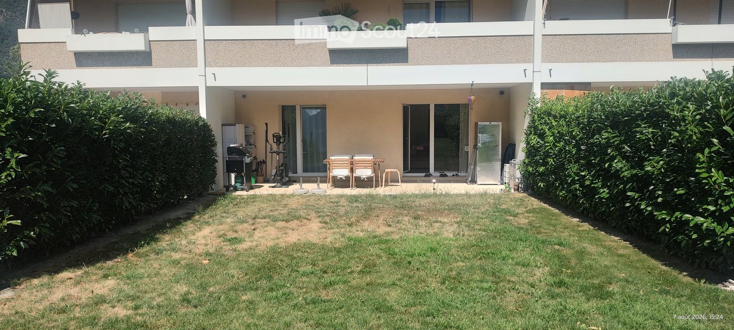 Well-maintained garden with a small patio area featuring outdoor furniture, a grill, and exercise equipment, surrounded by neatly trimmed hedges and a multi-story building in the background.
