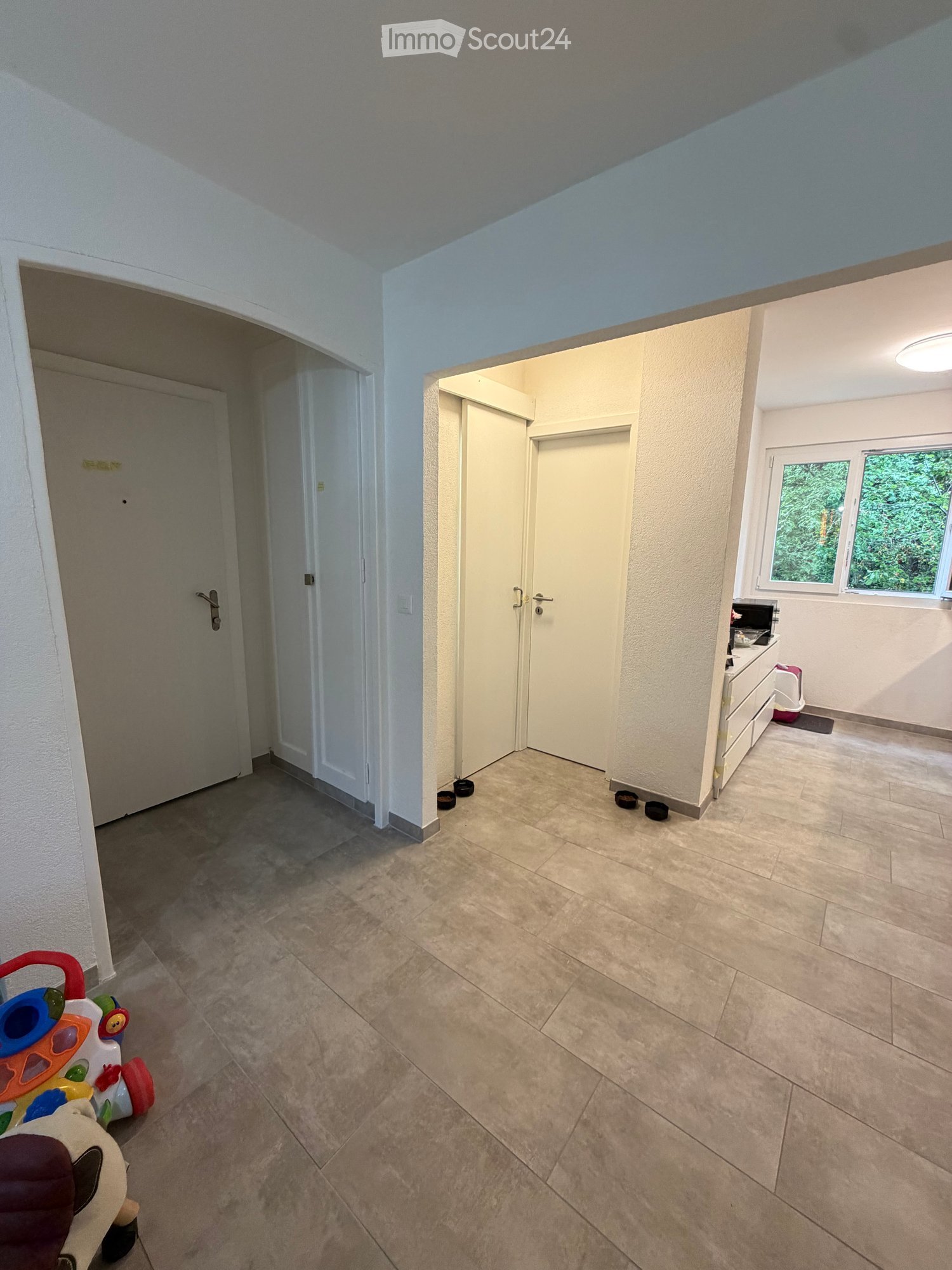 The image shows a hallway with tiled flooring and white walls. There is a toy stroller on the left side and a small stool near the entrance to another room. The hallway leads to a room with a window and a small table with a laptop on it.