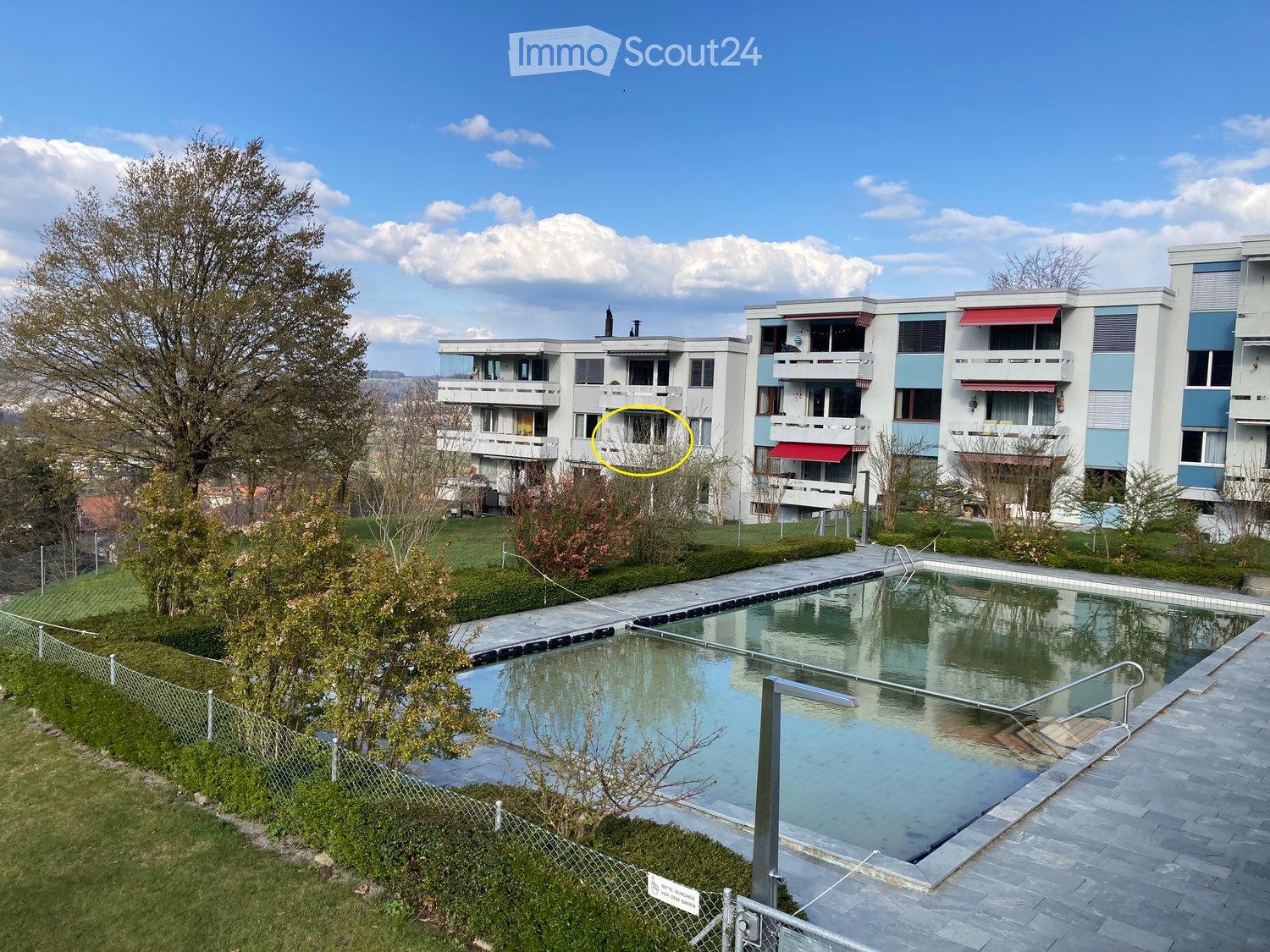 Outdoor area with a swimming pool, surrounded by a paved walkway, grassy areas, and trees. Modern apartment buildings with balconies and awnings are visible in the background.