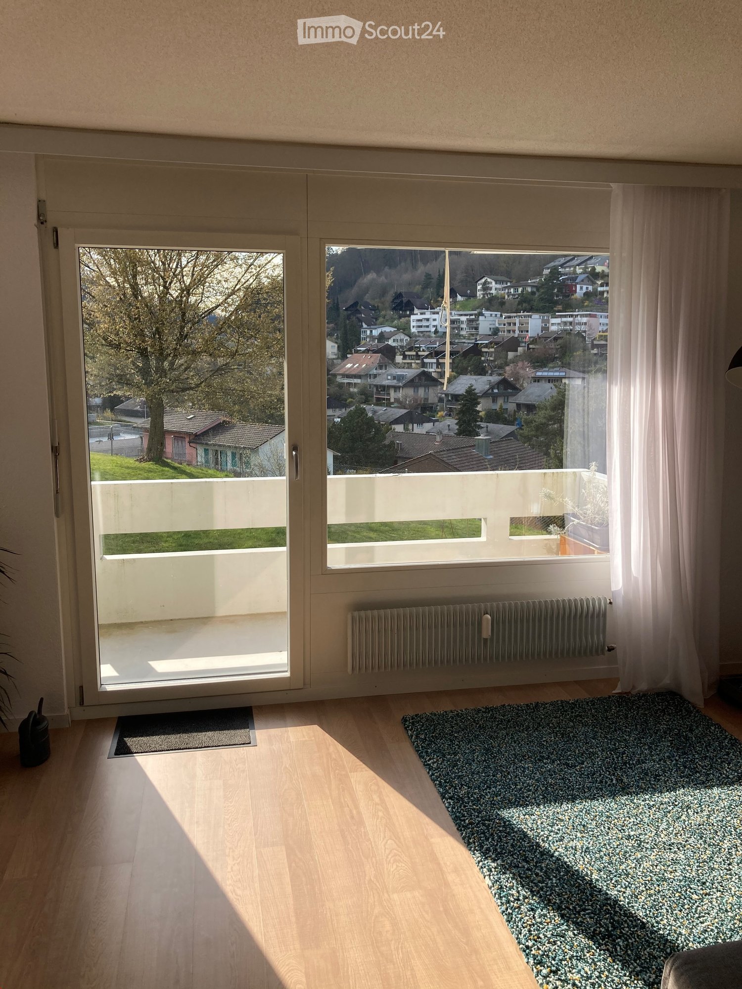 Bright room with wooden flooring, large windows offering a scenic view of a town and greenery, a patterned rug, and a radiator below the windows.