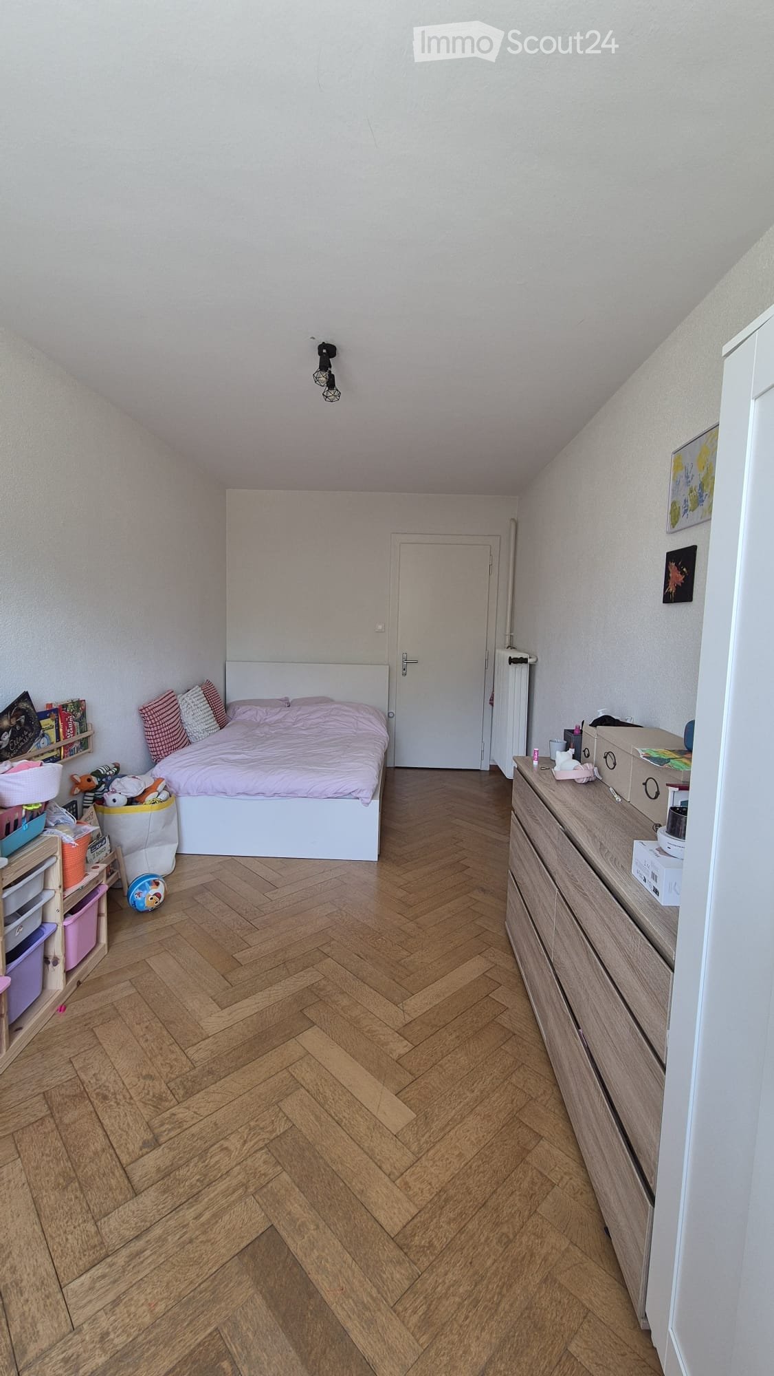 A small, cozy bedroom with wooden herringbone flooring, a white bed with pink bedding, a nightstand, a dresser, and a shelf with toys and books.