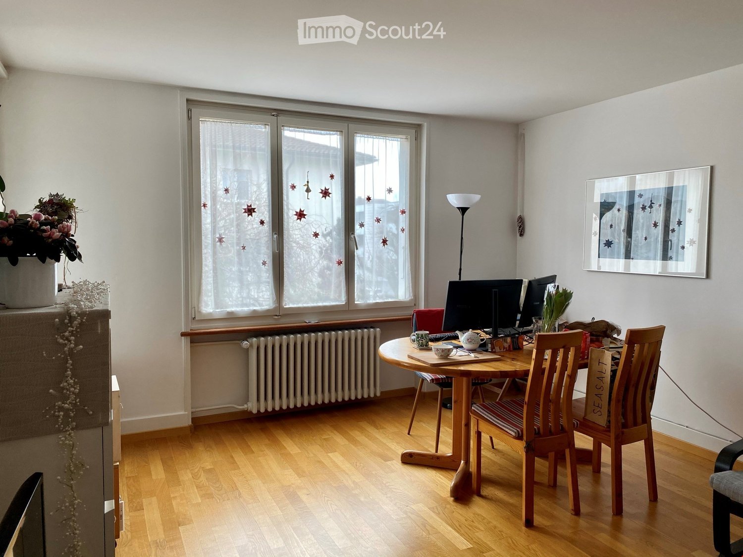 Bright room with wooden flooring, large window with decorative glass, round wooden dining table with chairs, computer setup, teapot and cups, flower pot, and picture frame on the wall.