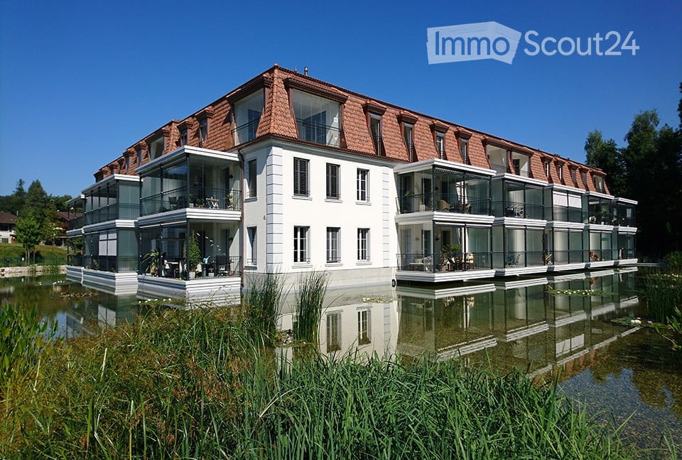 Modern two-story building with white walls, red-tiled roof, and large glass balconies overlooking a serene water body.
