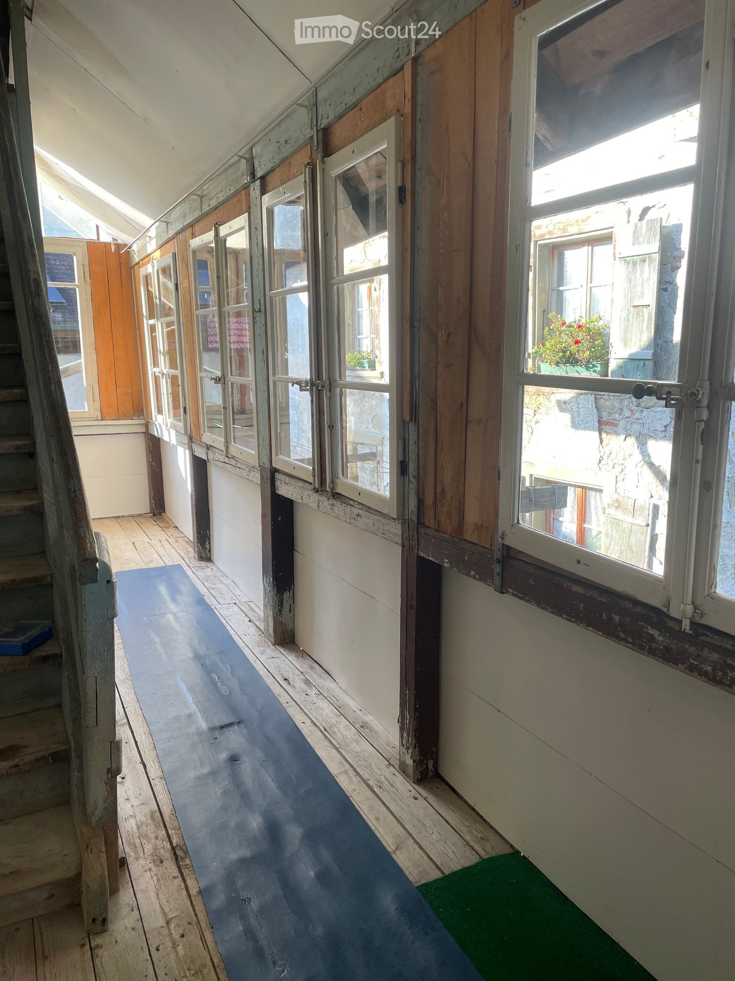 Empty hallway with wooden floors, white walls, and large windows with wooden frames, leading to a staircase on the left.